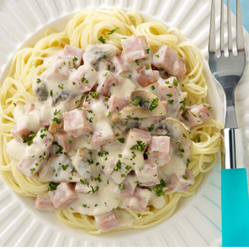 An easy, cheesy alfredo pasta packed with veggies and turkey ham, served on a white plate on a blue table.