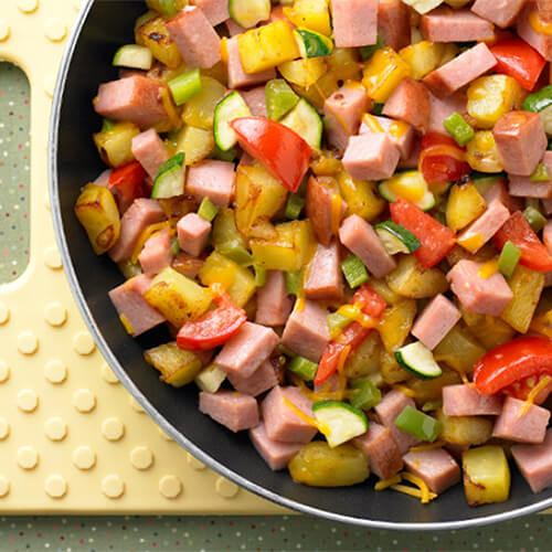 A skillet packed generously with a ton of fresh veggies, turkey ham, and melted Cheddar cheese, served on a yellow board.