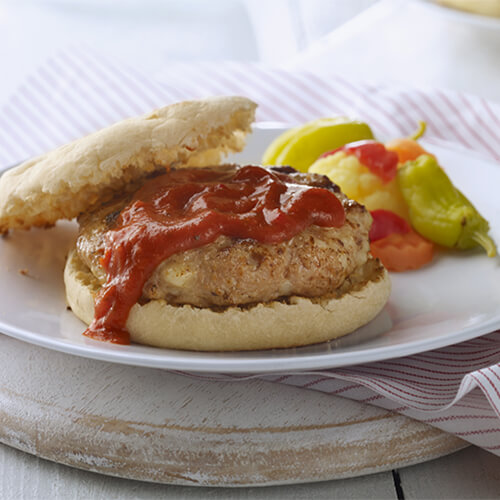 A flaky english muffin with pizza sauce and a patty that's a mixture of JENNIE-O® ground turkey, onion and mozzerella, served with peppers on the side, on a white plate, on top of a wooden platter.