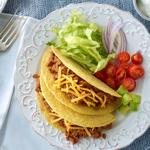 Turkey tacos made with cheese, lettuce, onion, and tomatoes, served on a white plate, on a blue woven tablecloth.