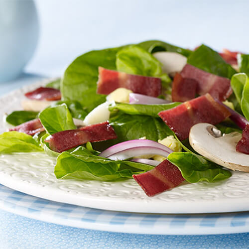 A hearty salad filled with vegetables, hard boiled eggs, mushrooms, and crispy turkey bacon, served in a white plate on a blue tablecloth.