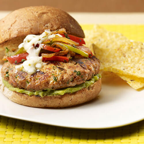 Spicy cumin-infused turkey patties, topped with Monterey jack cheese, guacamole and a hearty portion of sautéed bell peppers, served with a side of tortilla chips, on a white plate atop a yellow woven tablecloth.