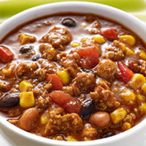 The tastes of red pepper, pinto beans, black beans, corn, green chilis, and tomatoes combine with the taste of turkey in a white bowl served with a side of celery on a white plate.