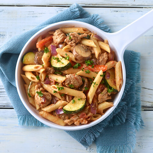 A skillet made with pasta, vegetables, ground turkey and CHI-CHI'S® salsa in a white soup bowl atop a blue napkin on a wooden background.