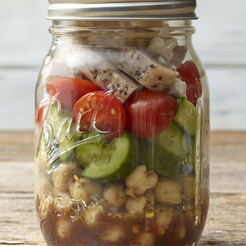 A balsamic vinaigrette, chickpeas, cucumber, tomatoes, turkey and feta cheese all served in a mason jar on a wooden table.