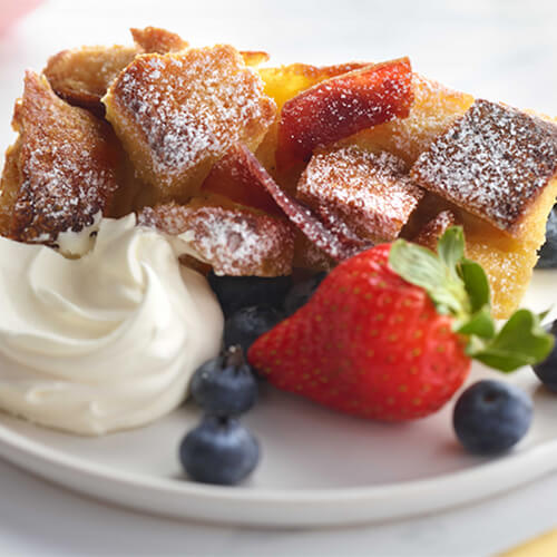 French toast bake with candied turkey bacon and fruits on a white plate.