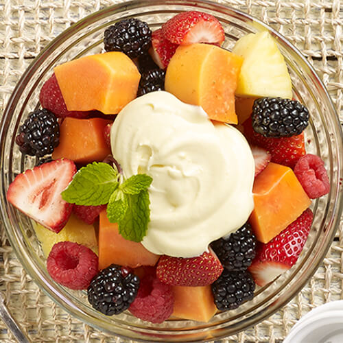 Fresh seasonal fruit with a dash of decadent cream made with hazelnut liqeur, served in a glass bowl on a wired tablecloth.