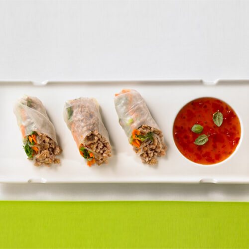 Spring rolls filled with cellophane noodles, a variety of fresh veggies, and ground turkey rolls, served with a side of sweet chili sauce on a white plate.