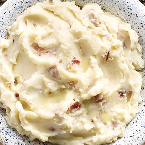 Garlic mashed potatoes made with turkey bacon and rosemary leaves, served on a painted bowl atop a wired table mat.