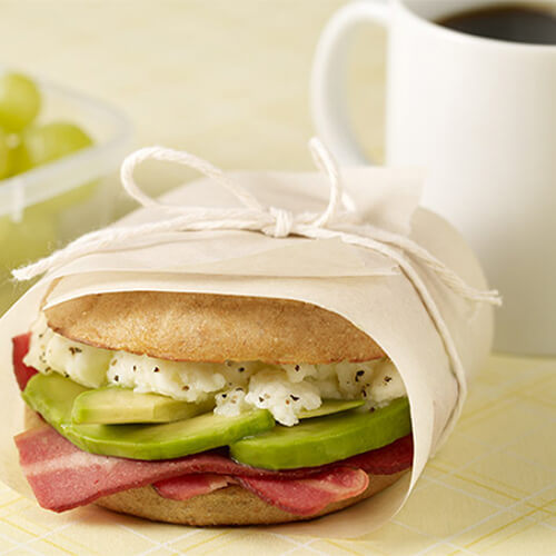 An English muffin, filled with bacon, avocado and scrambled egg white, wrapped in parchment paper, served with a cup of coffee and grapes.