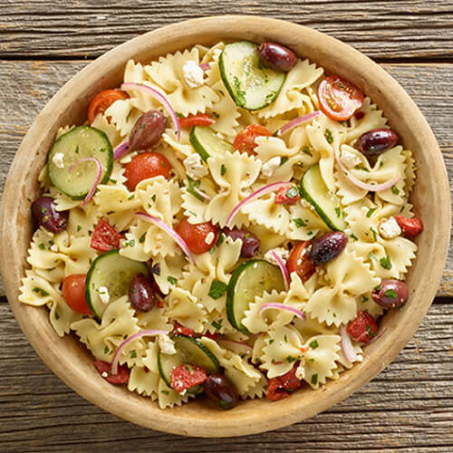 Cucumber slices, onion, olives, red bell pepper, tomatoes, herbs, and farfalle pasta served in a wood bowl garnished with feta, on a wooden table.