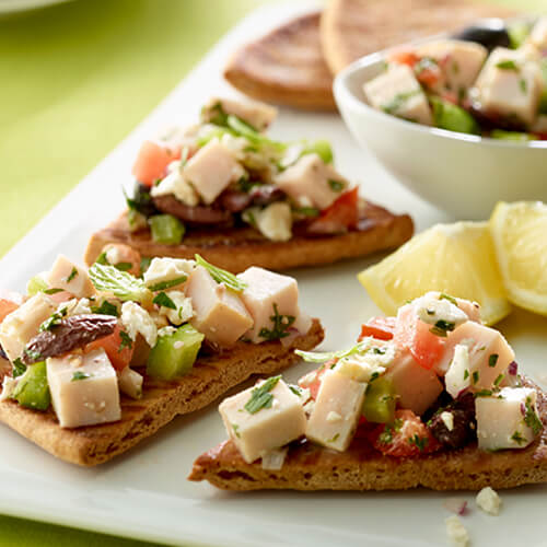 Wedges of pita bread, topped with turkey, olives, tomato, and parsley, served on a white platter with lemon wedges.