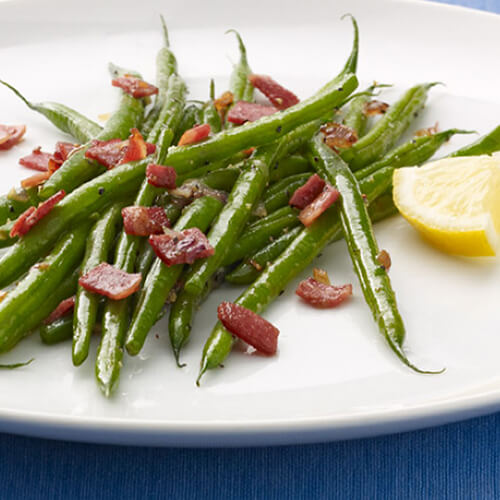 Tender-crisp green beans, sauteed with garlic, and topped with diced turkey bacon, served with a small lemon slice, on a white plate, atop a blue linen tablecloth.