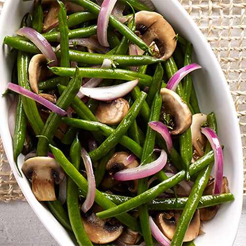 Crisp green beans and sautéed mushrooms topped with thinly sliced red onions, served in a white casserole tray on a wired table mat.