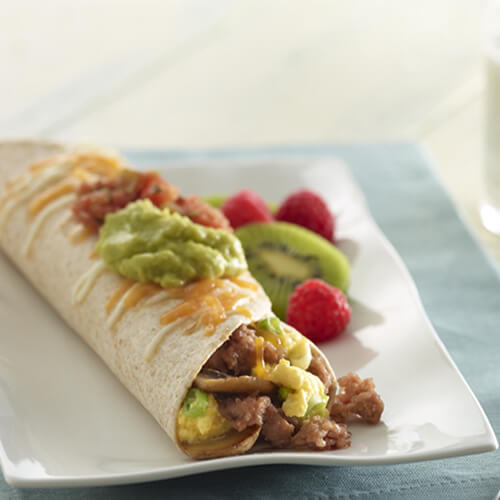 An irresistible burrito filled with lean turkey sausage, crispy turkey bacon and fluffy scrambled eggs, topped with melted cheese and guacamole, on a white plate, served with a side of fruit on a blue cloth.