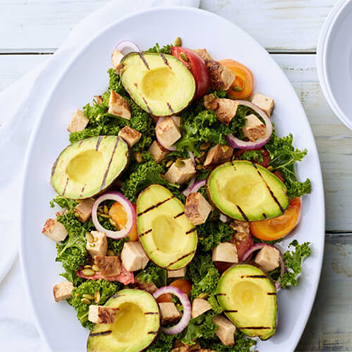 Grilled avocado and applewood turkey kale salad on a white oval dish on a white table.