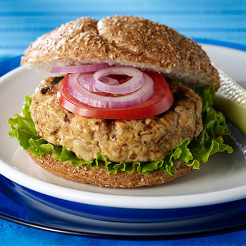 A burger patty made from turkey and beans, topped with onions, tomatoes, and lettuce, served on a whole-grain bun, with a white and blue plate garnished with dill pickles and a miniature tomato, atop a blue tablecloth.