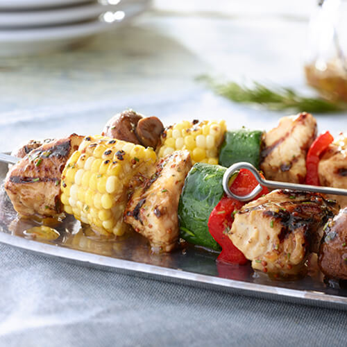 Kabobs made from mushrooms, zucchini, bell peppers, corn, and turkey tenderloins drizzled with a marmalade glaze on a metal plate on a gray tablecloth.