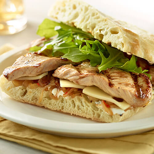 A grilled sandwich, featuring tender turkey breast, chili-balsamic onions, arugula, ranch dressing and Swiss cheese, served on a white plate atop a yellow napkin.