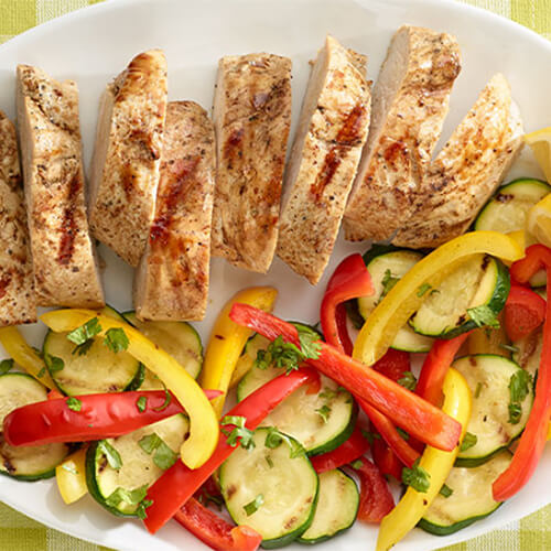 Cut turkey tenderloin, served with a hearty portion of grilled zucchini and bell peppers on a white plate atop a green tablecloth.