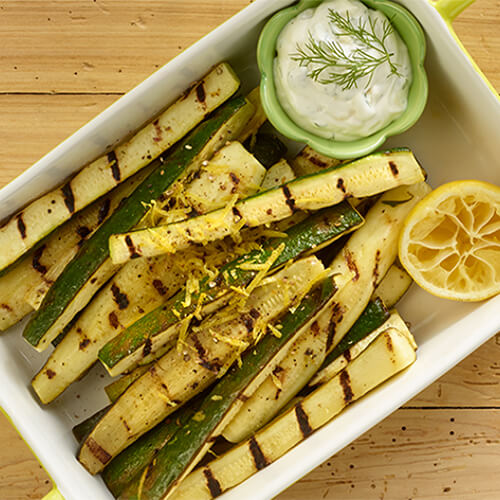 Grilled zucchini strips, served with a garlic mayo in a white tray atop a wooden table.