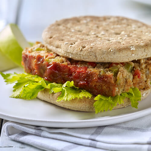 Sandwich thins topped with a celery leaves, mustard, and a meatloaf chock full of vegetables, served with a side of green apple on a white plate, on a wooden table.