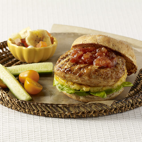 A turkey burger with grilled pineapple, lettuce, and topped with a combination of pineapple preserves and barbecue sauce, in a woven plate with a side of vegetables in a white background.