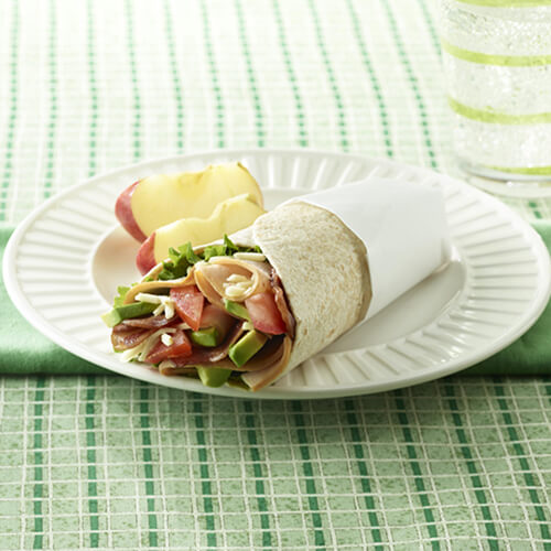 A wrap filled with fresh-sliced hickory-smoked turkey, bacon, and packed with vegetables, serves with a side of apples and sparkling water, on a white plate, atop green cloths.