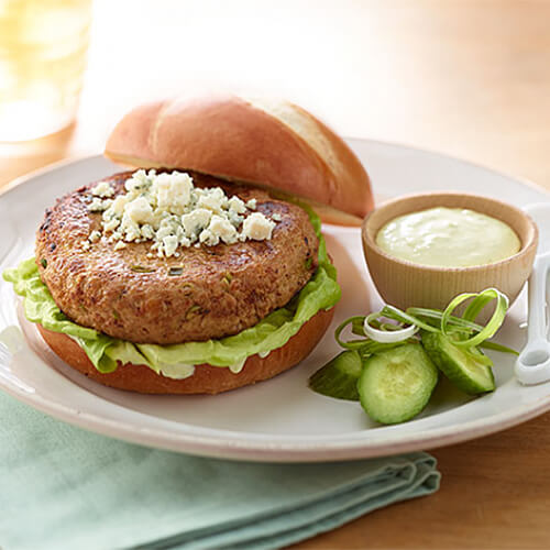 A spicy, breaded, turkey burger, topped with lettuce, crumbled blue cheese and Dijon on a whole wheat bun, served with jalapeno slices and a jalapeno mayo, on a white plate atop a blue cloth.