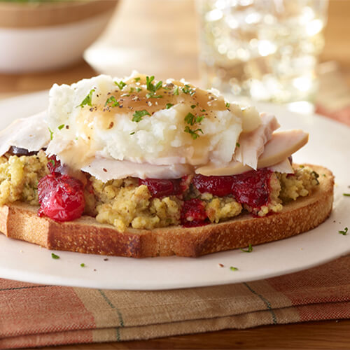 An open-face sourdough sandwich topped with stuffing, cranberry sauce, lean turkey, mashed potatoes and gravy, served on a white plate, garnished with parsley.