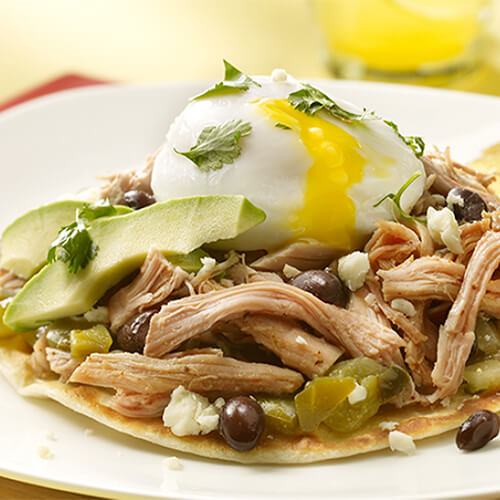 Open faced tacos filled with turkey pot roast, fresh avocado, cilantro, salsa, beans, and queso fresco, garnished with lime wedges on a white plate.