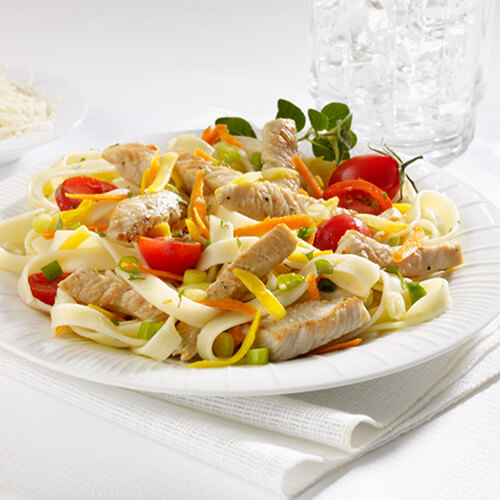 A hearty portion of fettuccine made with lean turkey tenderloins, and plenty of fresh veggies, and a white wine sauce, served on a white place with a side of parmesan cheese.
