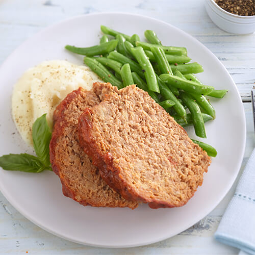 Two thick slices of meatloaf over a side of mashed potatoes and seasoned green beans on a white plate.