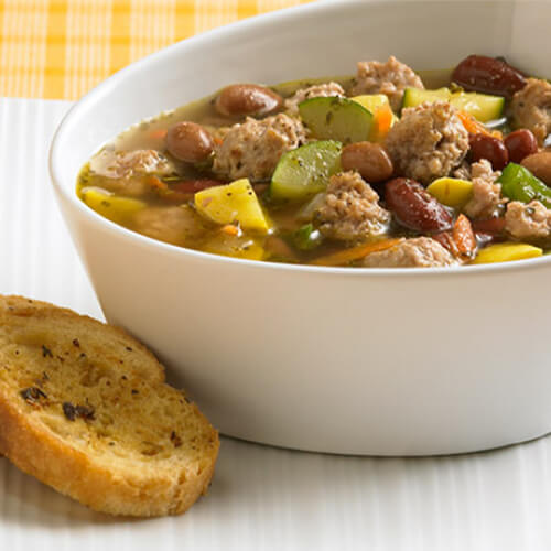 A hearty portion of minestrone soup filled with ground turkey breast, vegetables, beans and Herb-Ox bouillions in a white bowl, with a side of crackers, on a white plate, in a yellow flannel tablecloth.