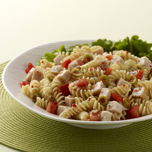 Rotini pasta topped with a hearty portion of smoked turkey breast, bell peppers, and with a side of salad greens, on a white bowl, on a green placemat and background.