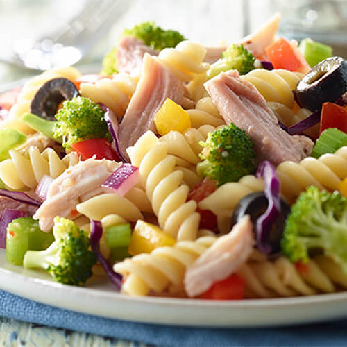 Turkey salad made with cooked pasta, onion, olives, peppers, tomato, and more vegetables, tossed with turkey and an Italian salad dressing on a white plate atop a blue cloth.