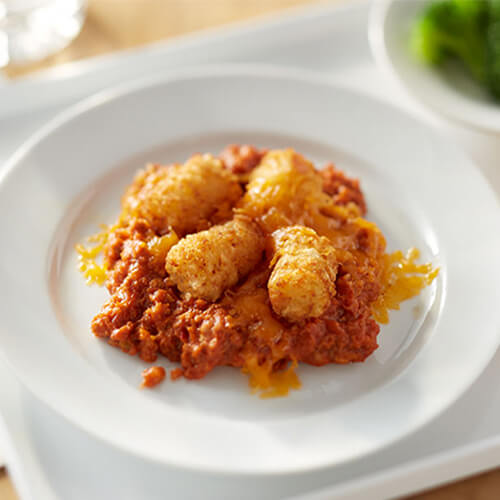 Turkey sprinkle with cheese and topped with marinara sauce and tater tots, served on a white plate with a side of broccoli and fruit on a white tray.