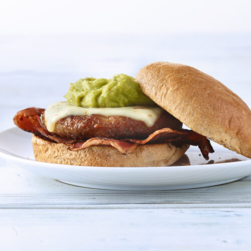 An amazing turkey burger topped with bacon strips, guacamole and melted pepper jack cheese. served on a white plate on a wooden table.