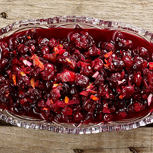 A sweet cranberry sauce made with honey, lime leaves and cinnamon sticks, served in a glass dish atop a wooden table.