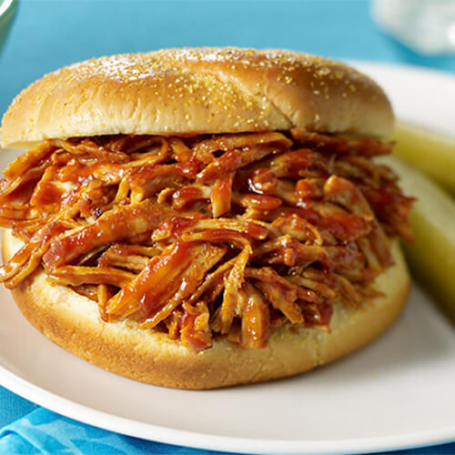 A hearty portion of barbecue sauce covered turkey, served on a white plate with a side of pickles and coleslaw, on a cyan tablecloth.