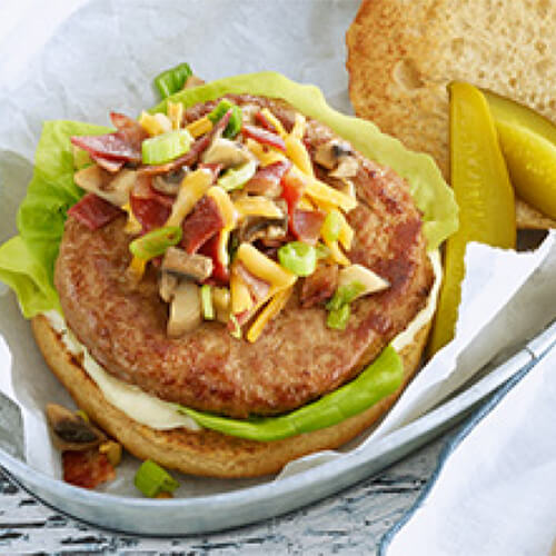 A turkey burger loaded with turkey bacon, cheese, mushrooms, onion, mayonaise, and lettuce, served on a hamburger bun in a metal tray with a side of chopped pickles.