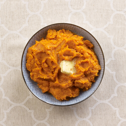 Mashed sweet potatoes garnished with a pad of butter in a blue bowl on a beige patterned tablecloth.