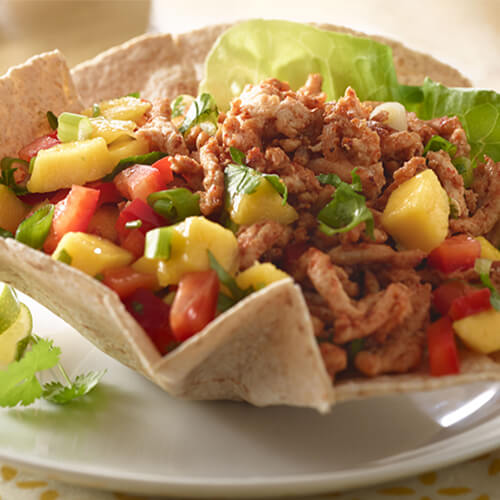 A hearty taco salad filled fresh mango salsa, veggies, and seasoned ground turkey, topped with cilantro, garnished with cilantro and lime wedges served on a white plate.