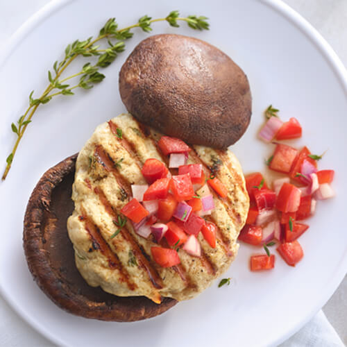 A hearty turkey burger topped with fresh veggies, a Portobello mushroom cap, and with a side of Mediterranean yogurt sauce, on a white plate and table.