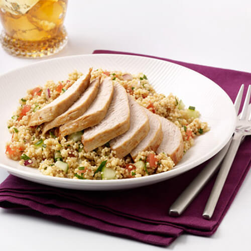 A healthy amount of mediterranean-flavored turkey on a bed of soft couscous, tossed with fresh vegetables on a white plate on a mauve napkin.