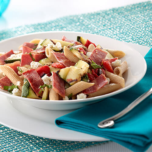 Crumbled feta, green olives and grilled zucchini join whole wheat pasta topped with a zesty lemon garlic dressing, served on a white plate and bowl atop a blue woven table mat.