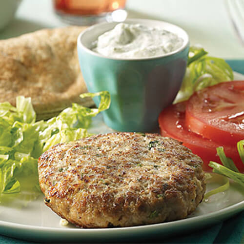 A Mediterranean-style turkey burger served with lettuce, tomato, pitas and tzatziki, served on a gray plate atop a cyan cloth.