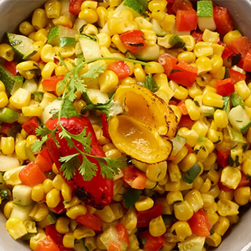 Juicy kernels of corn sautéed with red pepper, zucchini, green onion and jalapeno, served in a white bowl atop a gray tablecloth.