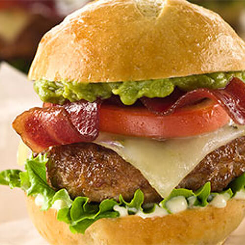 A miniature turkey burger filled with lettuce, tomato, bacon, and cheese, fresh guacamole, and green chili mayo, served on parchment paper.