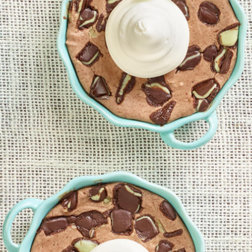 Decadent mint chocolate cheesecakes made with crushed cookie crumbs and chocolate mint candies, served in blue custard cups topped with a dollop of whipped cream, served on a linen tablecloth.